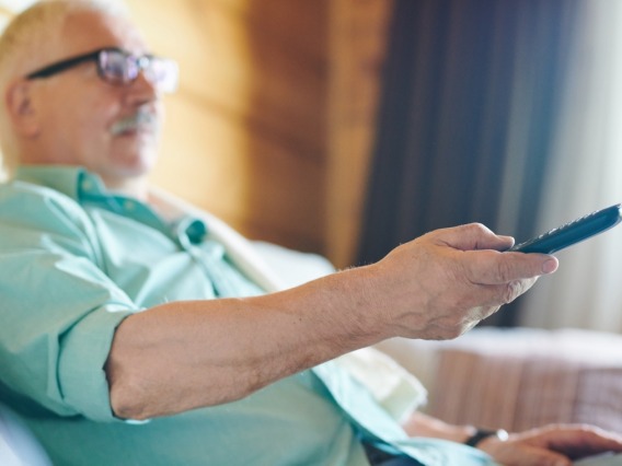 Older white male with glasses and mustache sitting with TV television remote in chair during afternoon daytime.