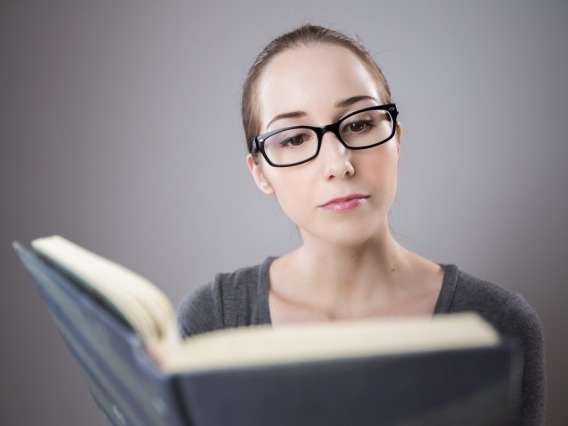 Caucasian female with glasses serious-faced and reading an open book in front of grey background