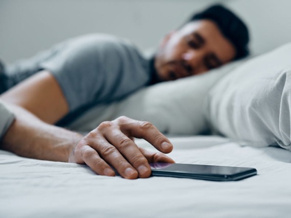 Latino man laying in bed with grey sheets and reaching for cell phone