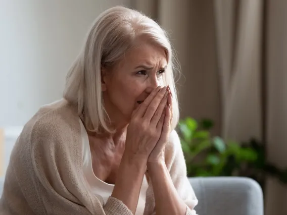 Elder white woman with white hair holding hands to face in tan sweater on sofa with daylight coming through window