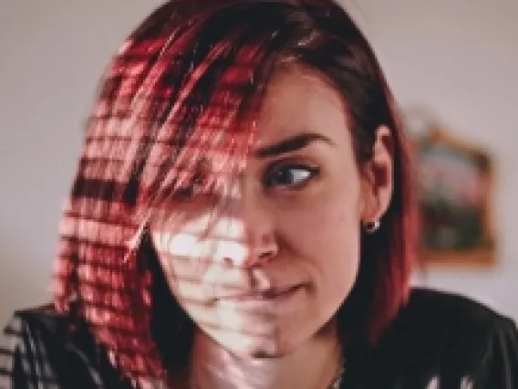 Caucasian girl with red-dyed hair pensively looking to left side with tight lips with sunlight coming across face through window blinds