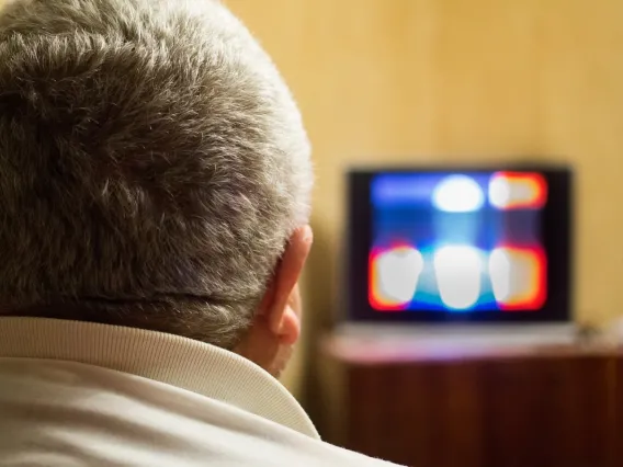 Back of head of male with short grey hair in foreground with blurry television screen in background