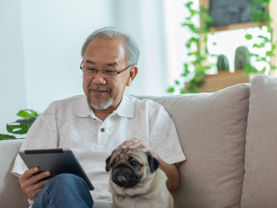 Older Asian man with glasses in white button-up shirt reading his iPad on beige couch with pug dog sitting beside him