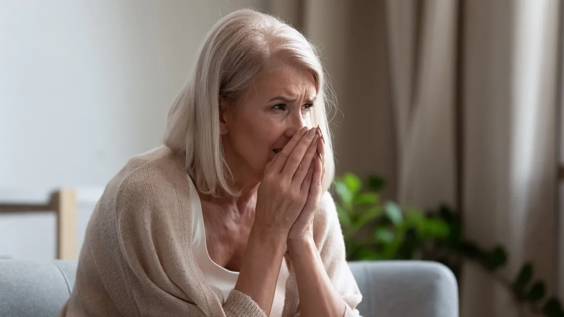 Elder white woman with white hair holding hands to face in tan sweater on sofa with daylight coming through window