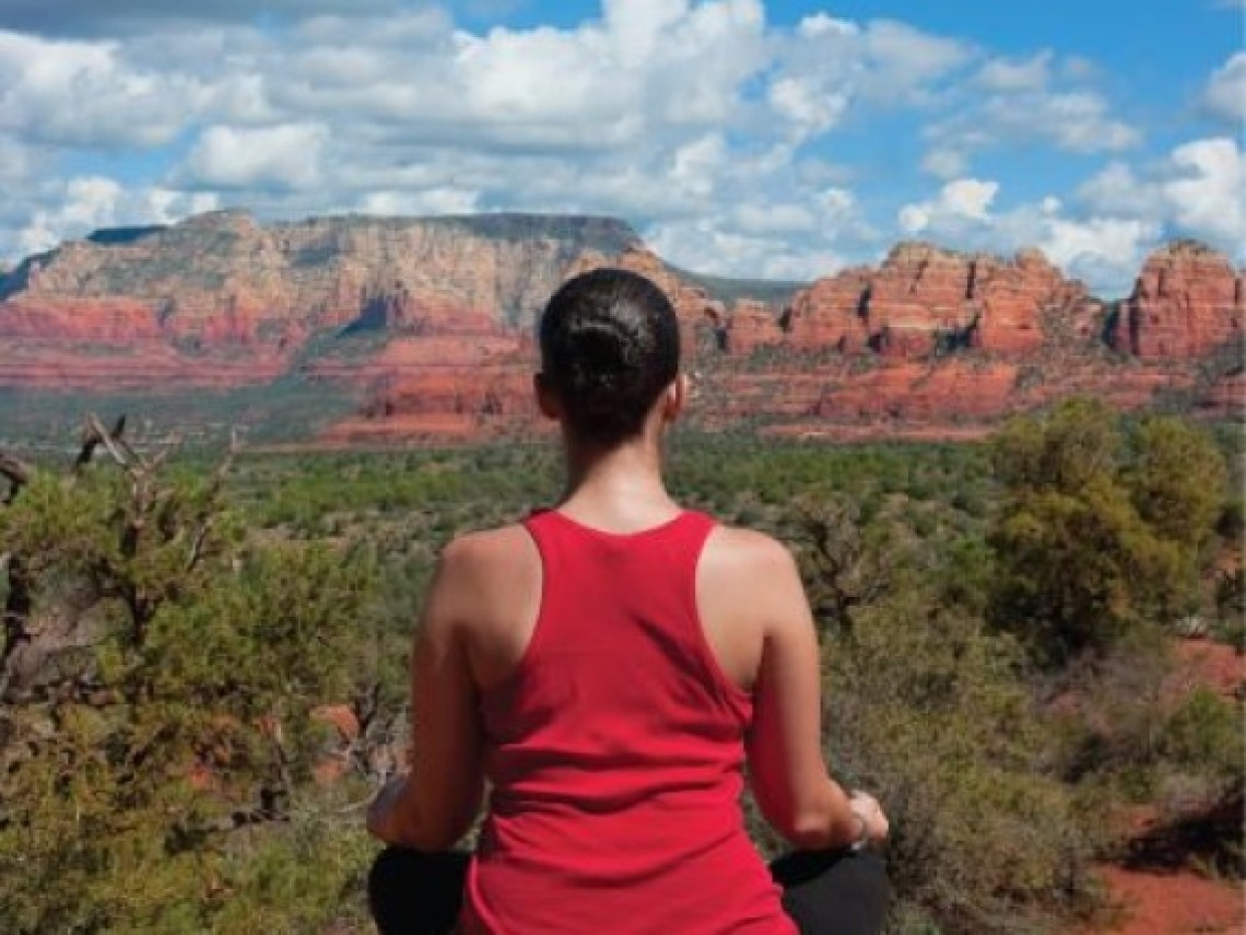 Cacausian brunette woman in red tank top with back turned meditating while facing red rock mesa under the clouds