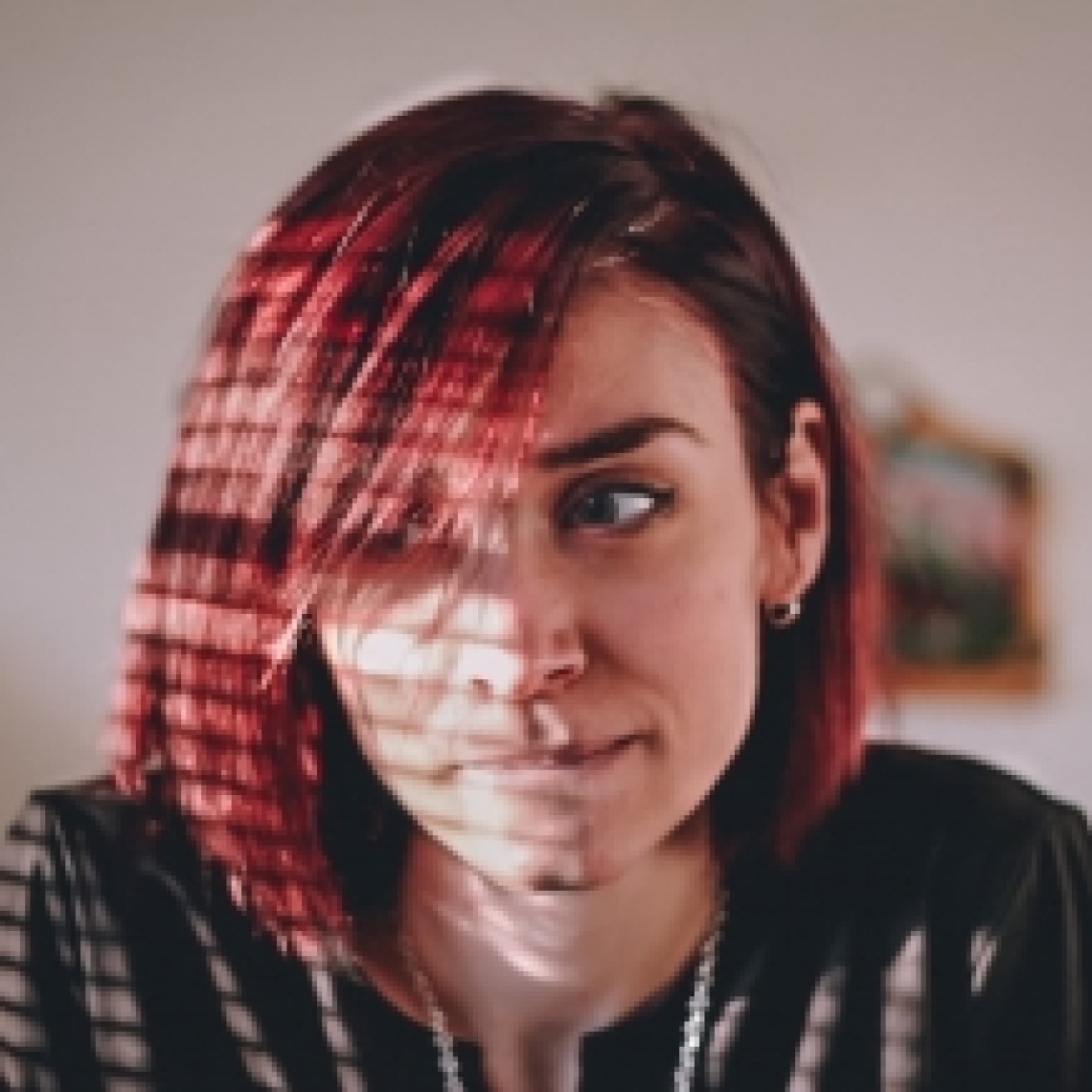 Caucasian girl with red-dyed hair pensively looking to left side with tight lips with sunlight coming across face through window blinds