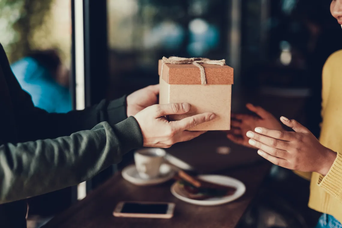 Caucasian male in dark green sweater giving brown gift box to African American woman in yellow sweater