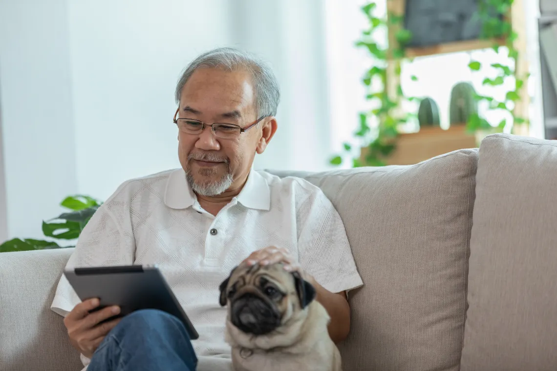 Older Asian man with glasses in white button-up shirt reading his iPad on beige couch with pug dog sitting beside him