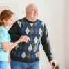 Auburn-haired lady in teal T-shirt and jeans taking arm of older bald man in argyle sweater & jeans with cane, assisting him to walk