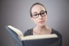 Caucasian female with glasses serious-faced and reading an open book in front of grey background