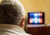 Back of head of male with short grey hair in foreground with blurry television screen in background